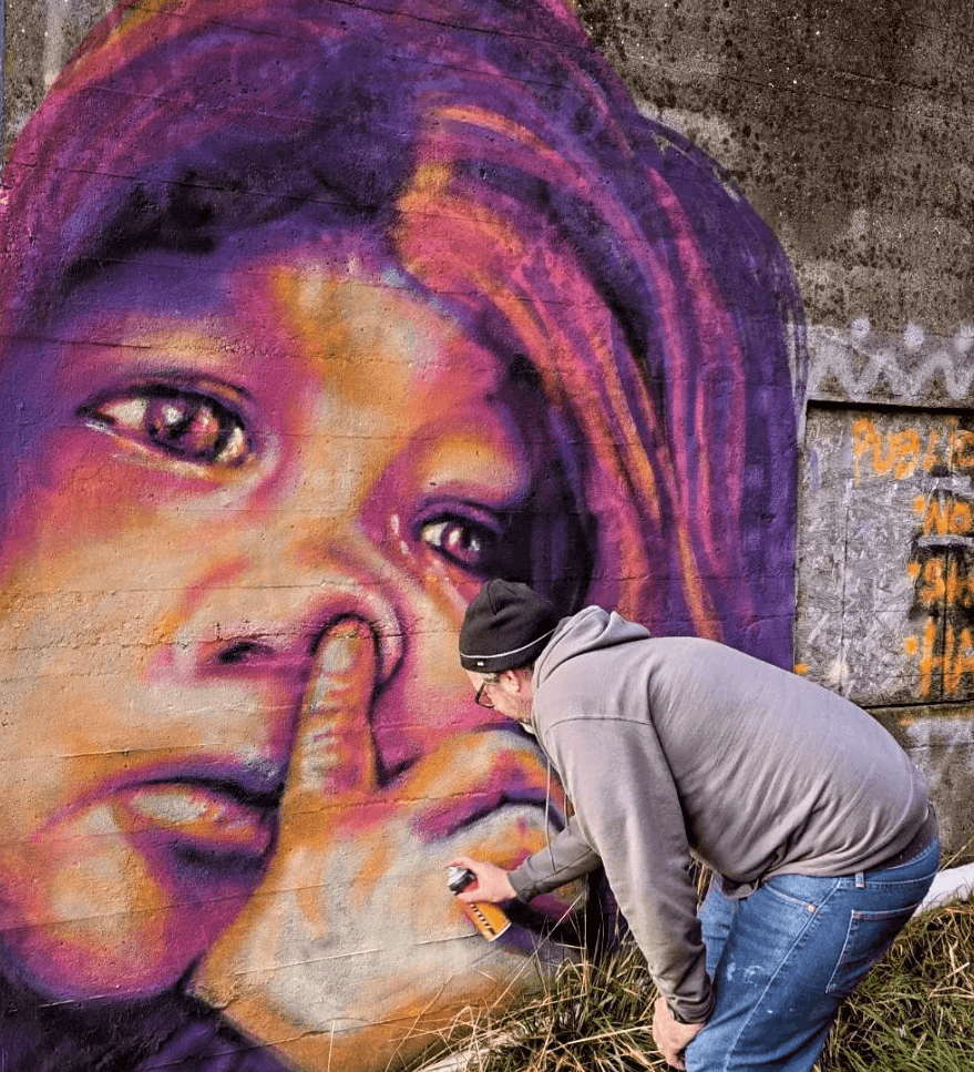 Artist Brady Black with a spray paint can doing art on the public art wall of The Hub in downtown Port Angeles Washington. Art is a mural of a young girl picking her nose looking up to the right corner featuring vibrant colors.
